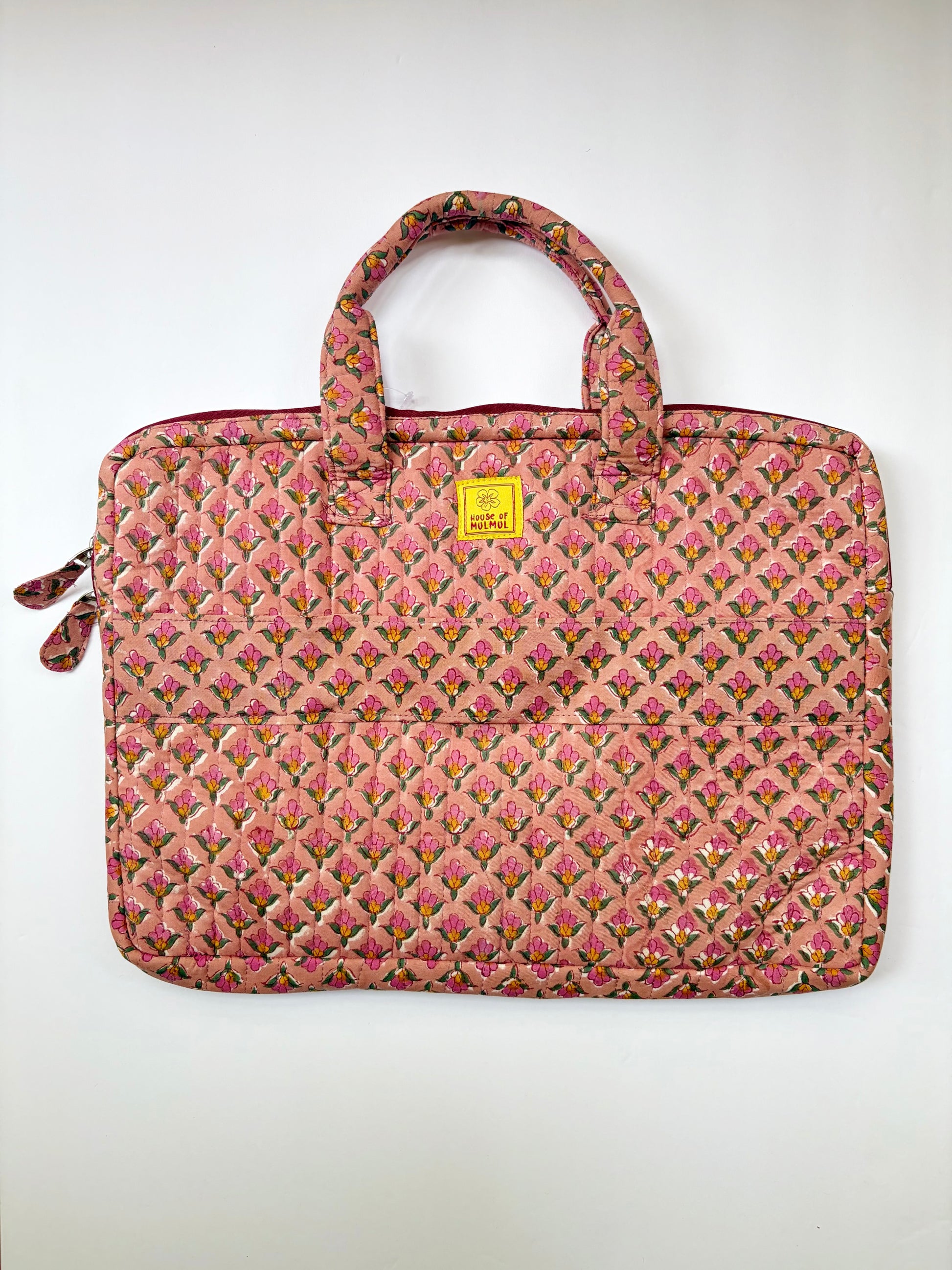 House of Mulmul - Blockprinted Cotton Quilted Laptop Bag. A quilted pink laptop sleeve featuring a repeating floral pattern in shades of pink, green, and yellow, with dual top handles and a small yellow brand tag on the front.