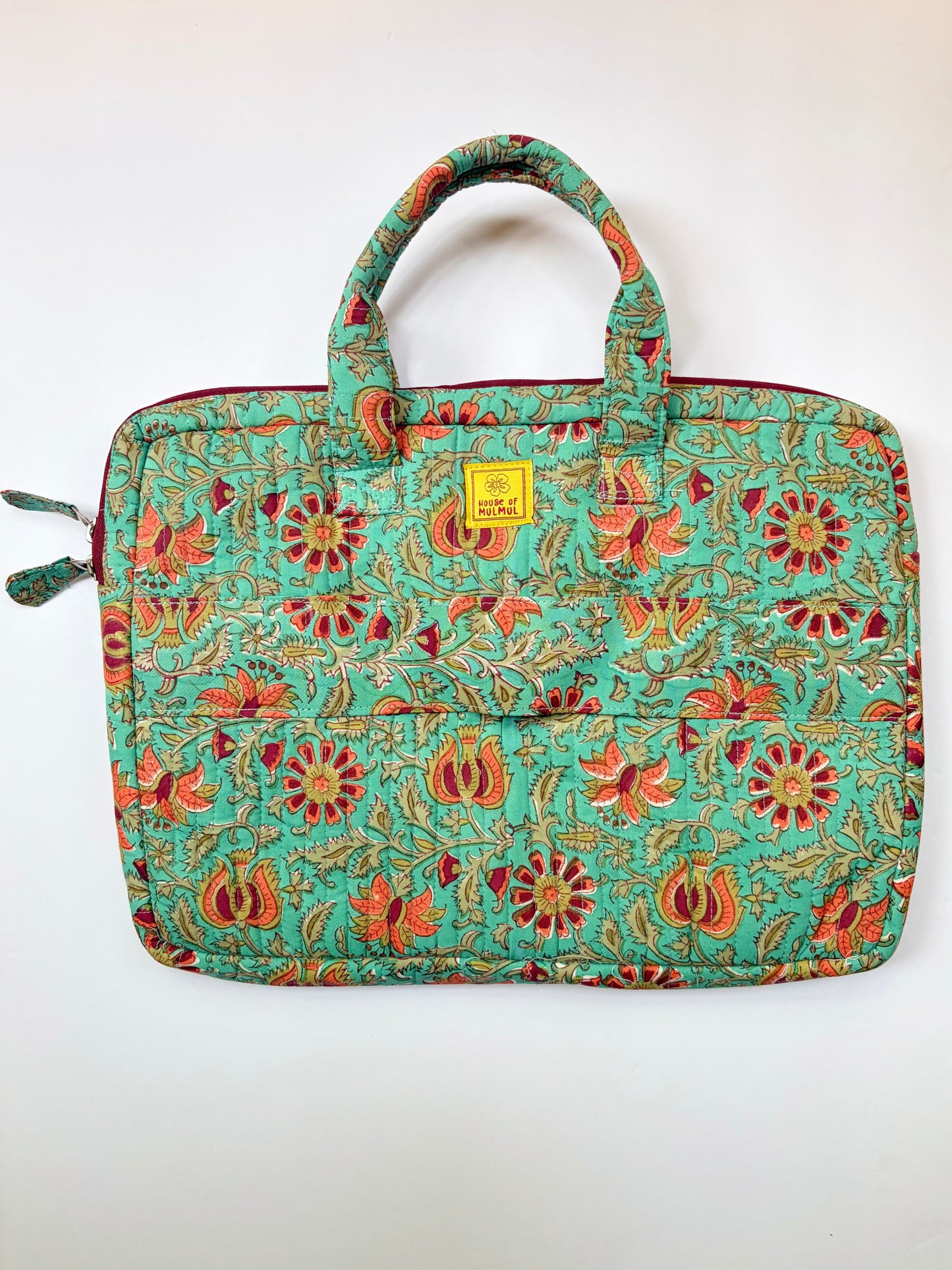 House of Mulmul - Blockprinted Cotton Quilted Laptop Bag. A quilted laptop sleeve featuring a vibrant floral and paisley pattern in turquoise, red, orange, and gold. It has two top handles, a zippered closure, and a small yellow brand label on the front.