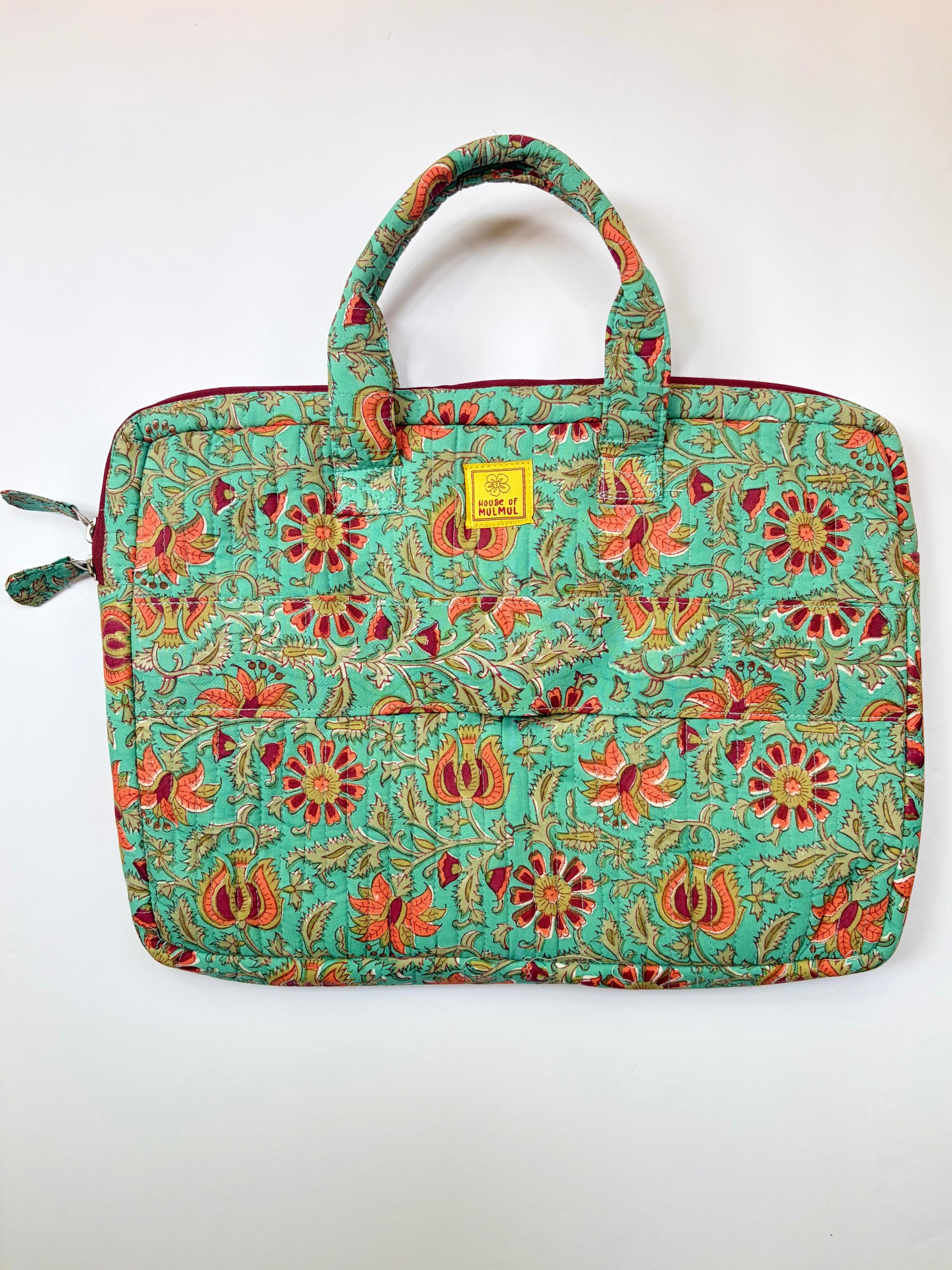 House of Mulmul - Blockprinted Cotton Quilted Laptop Bag. A quilted laptop sleeve featuring a vibrant floral and paisley pattern in turquoise, red, orange, and gold. It has two top handles, a zippered closure, and a small yellow brand label on the front.