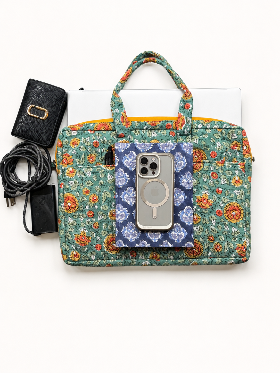 House of Mulmul - Blockprinted Cotton Quilted Laptop Bag. A floral tote-style laptop bag with a green, orange, and blue pattern, shown flat-lay with a laptop, phone in a blue floral case, a black wallet, and a black power cable on a white background.