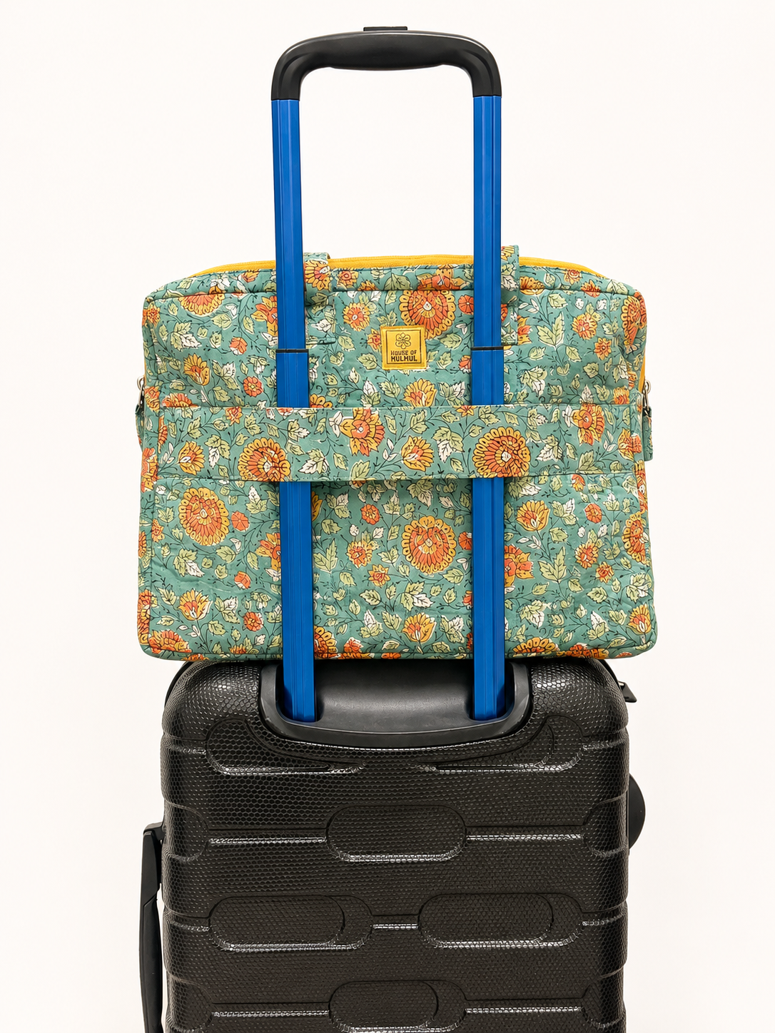 House of Mulmul - Blockprinted Cotton Quilted Laptop Bag. A floral carry-on laptop bag with a blue telescoping handle resting on top of a black hard-shell suitcase against a plain off-white background.