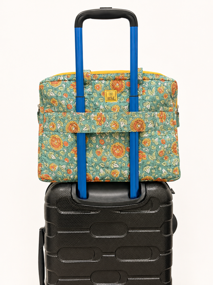 House of Mulmul - Blockprinted Cotton Quilted Laptop Bag. A floral carry-on laptop bag with a blue telescoping handle resting on top of a black hard-shell suitcase against a plain off-white background.