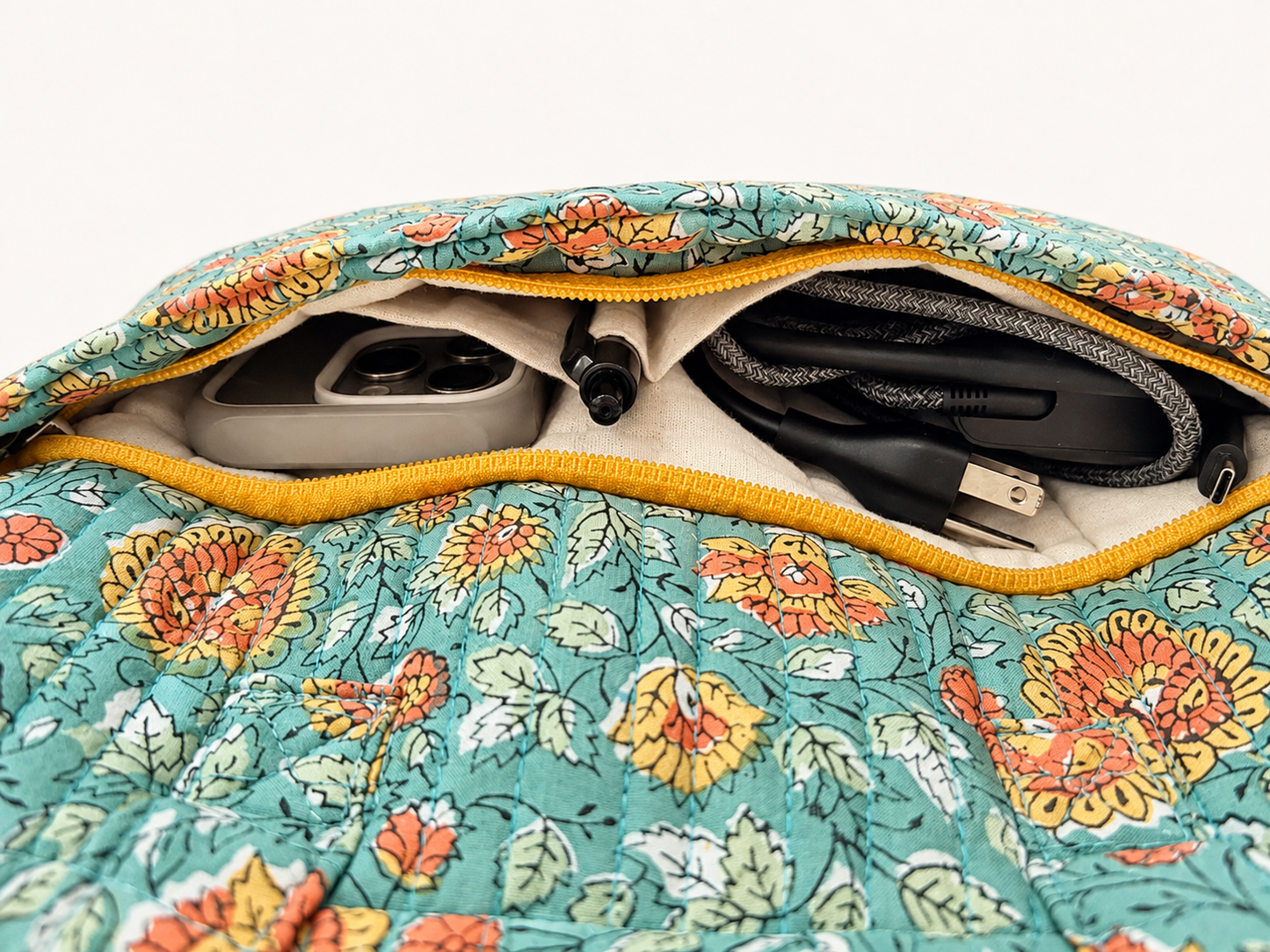 House of Mulmul - Blockprinted Cotton Quilted Laptop Bag. Close-up of a floral quilted laptop bag with a yellow zipper, showing its interior compartments containing a gray power bank, a black pen, and a gray cable.