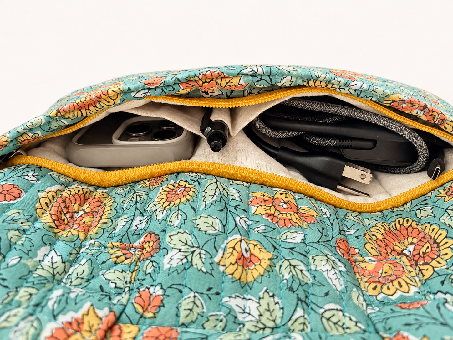 House of Mulmul - Blockprinted Cotton Quilted Laptop Bag. Close-up of a floral quilted laptop bag with a yellow zipper, showing its interior compartments containing a gray power bank, a black pen, and a gray cable.