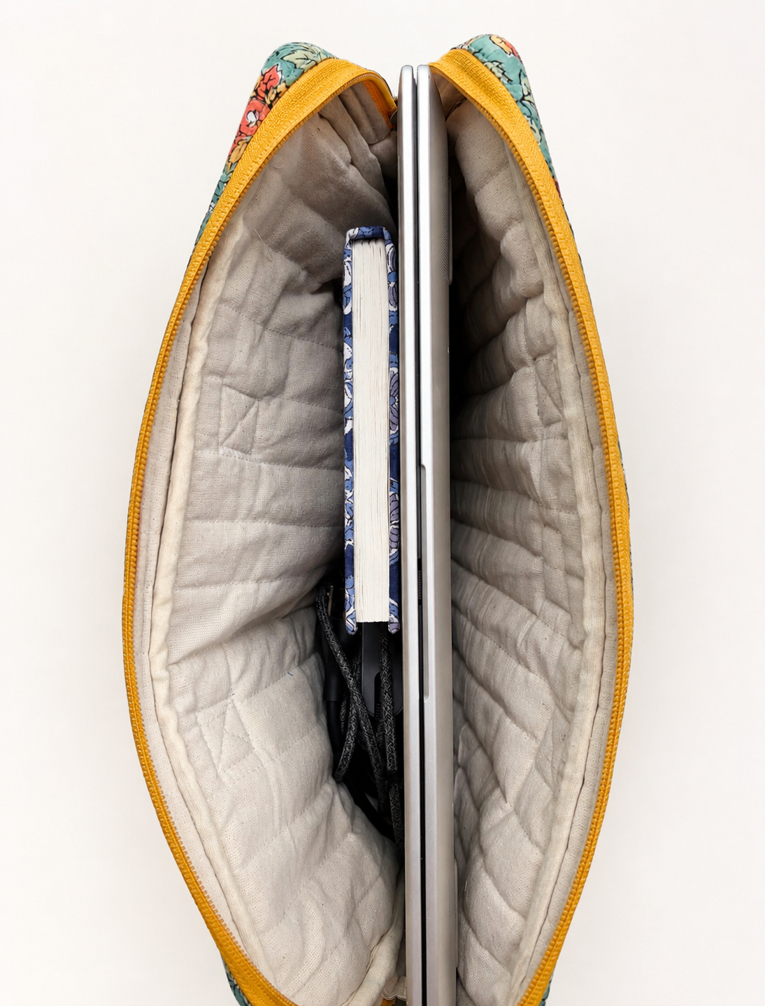 House of Mulmul - Blockprinted Cotton Quilted Laptop Bag. An open quilted laptop bag with a mustard yellow zipper, cream interior, and a multicolor floral fabric exterior, shown with a silver laptop and notebook inside on a white background.