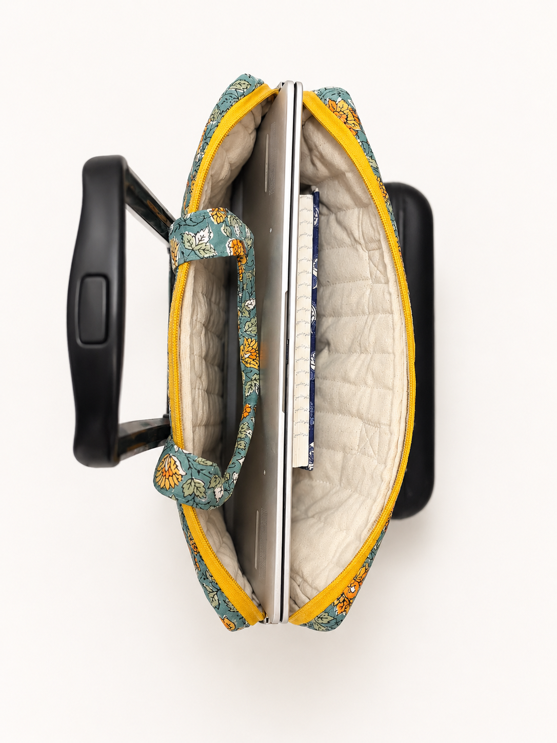 House of Mulmul - Blockprinted Cotton Quilted Laptop Bag. An open quilted laptop bag with a floral pattern in teal, yellow, and orange, shown from a top-down view with a black rolling suitcase handle inserted on either side.