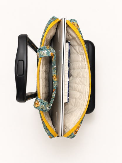 House of Mulmul - Blockprinted Cotton Quilted Laptop Bag. An open quilted laptop bag with a floral pattern in teal, yellow, and orange, shown from a top-down view with a black rolling suitcase handle inserted on either side.