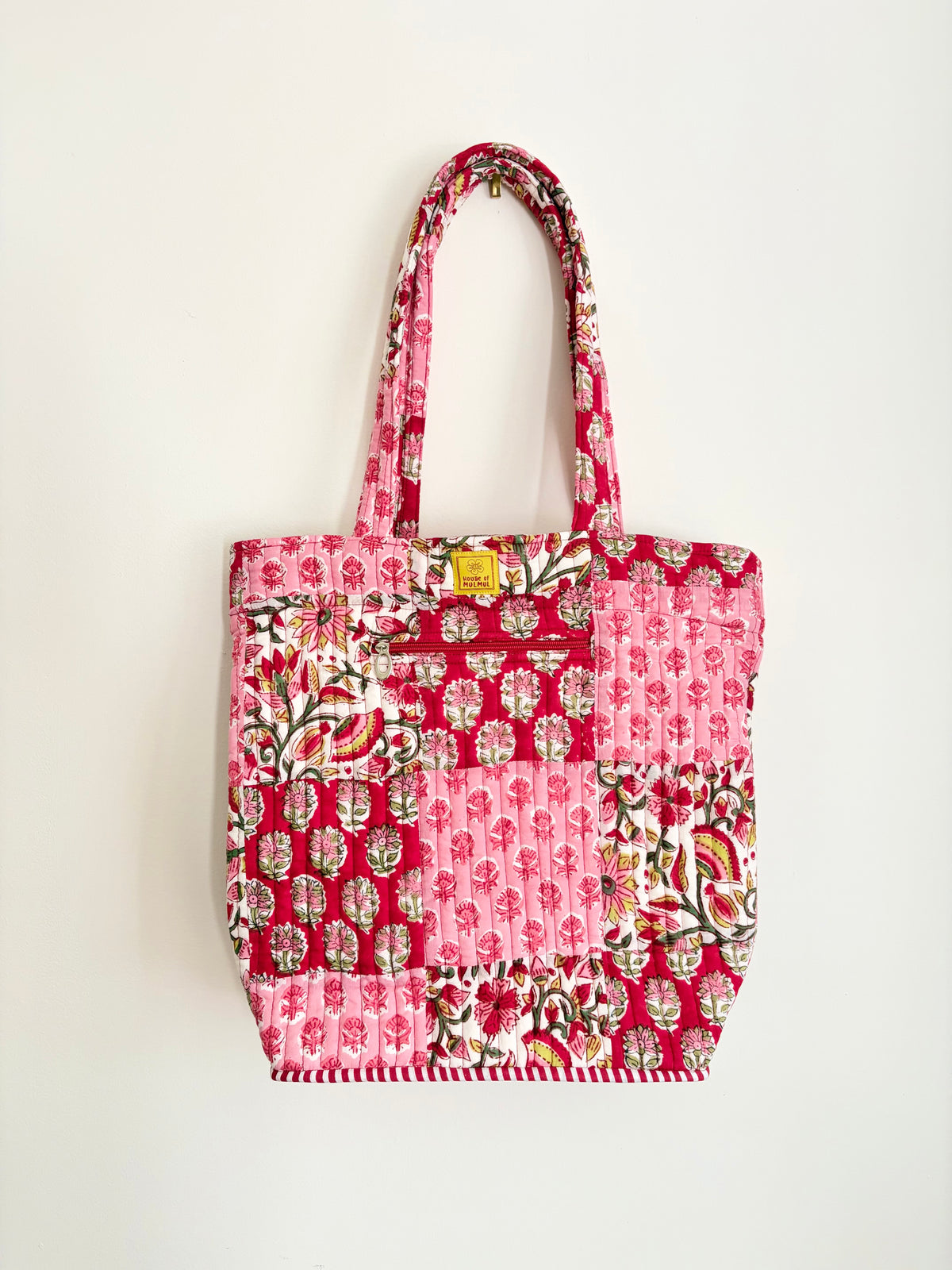 House of Mulmul - Bucket Tapered Tote Bag. A pink patchwork quilted fabric tote bag with floral and paisley patterns, featuring two top handles and a small yellow brand tag, shown against a plain white background.