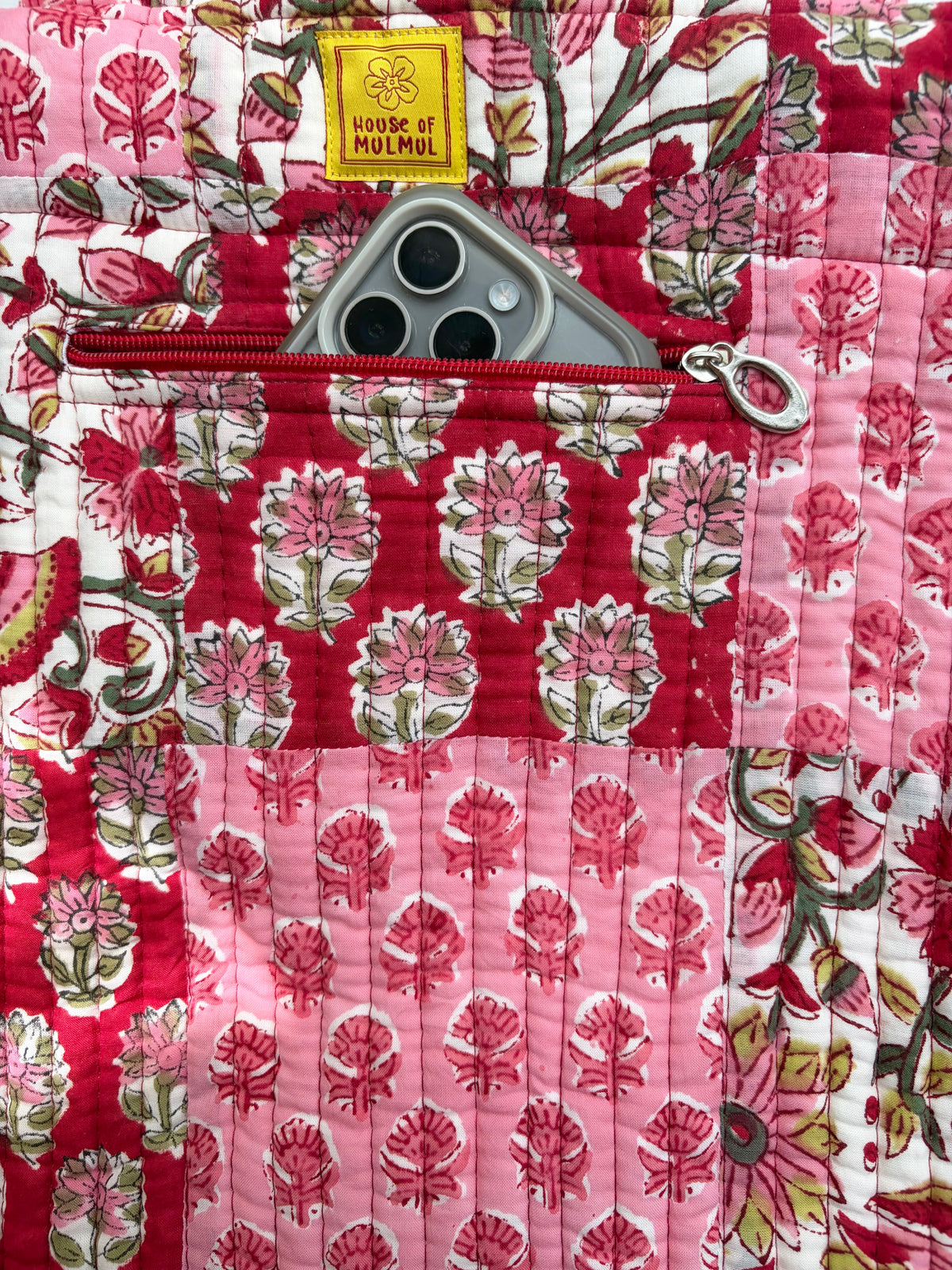 House of Mulmul - Bucket Tapered Tote Bag. A close-up of a quilted tote bag in a patchwork floral print, featuring a red pocket with a silver zipper and a gray smartphone inserted. A yellow fabric label reads 'House of Mulmul'.