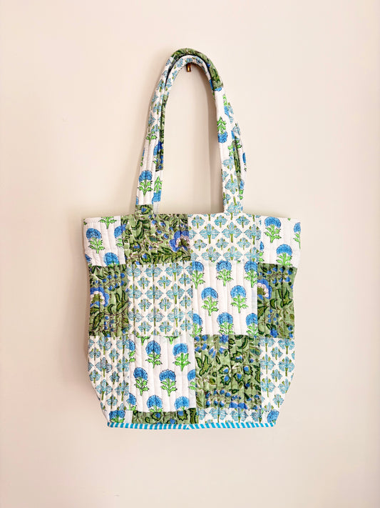 House of Mulmul - Bucket Tapered Tote Bag. A handmade patchwork tote bag featuring blue and green floral prints on white fabric panels, hanging against a solid beige wall.