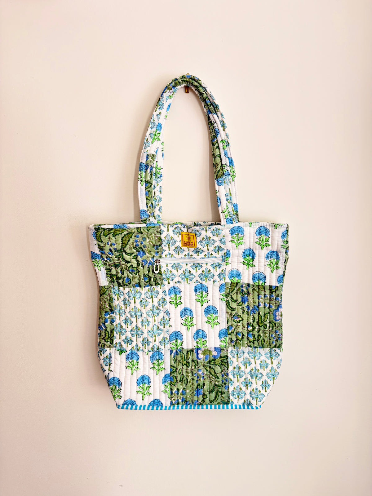 House of Mulmul - Bucket Tapered Tote Bag. A patchwork quilted tote bag with blue and green floral prints on a white background, featuring two shoulder straps and a small gold brand tag, hanging against a solid pale pink background.