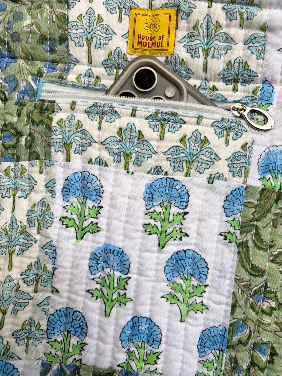 House of Mulmul - Bucket Tapered Tote Bag. Close-up of a quilted fabric pouch with a floral print in blue, green, and white, featuring a yellow 'House of Mulmul' brand label and a silver zipper.
