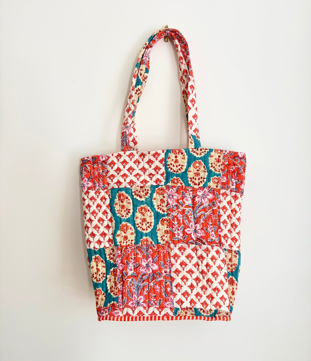 House of Mulmul - Bucket Tapered Tote Bag. A patchwork fabric tote bag with red, teal, and cream floral and geometric patterns, featuring two shoulder straps, shown hanging against a plain white wall.