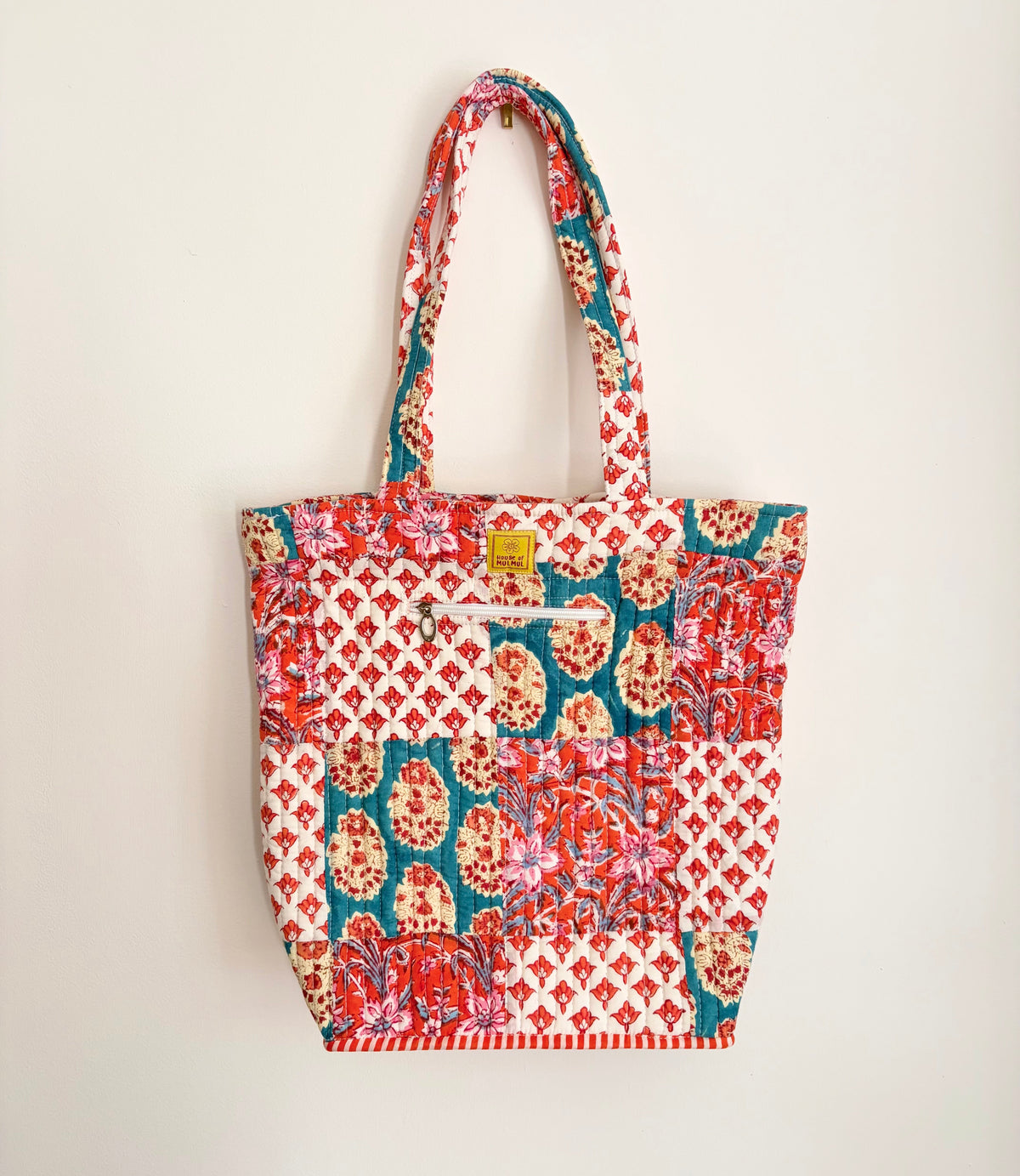 House of Mulmul - Bucket Tapered Tote Bag. A patchwork quilted tote bag with red, teal, and cream floral patterns, featuring two shoulder straps and a small gold metal logo on the front.