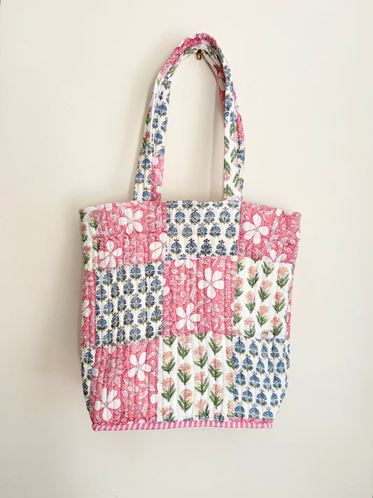 House of Mulmul - Bucket Tapered Tote Bag. A handmade patchwork quilted tote bag with floral and geometric patterns in pink, blue, and white, hanging against a plain off-white wall.