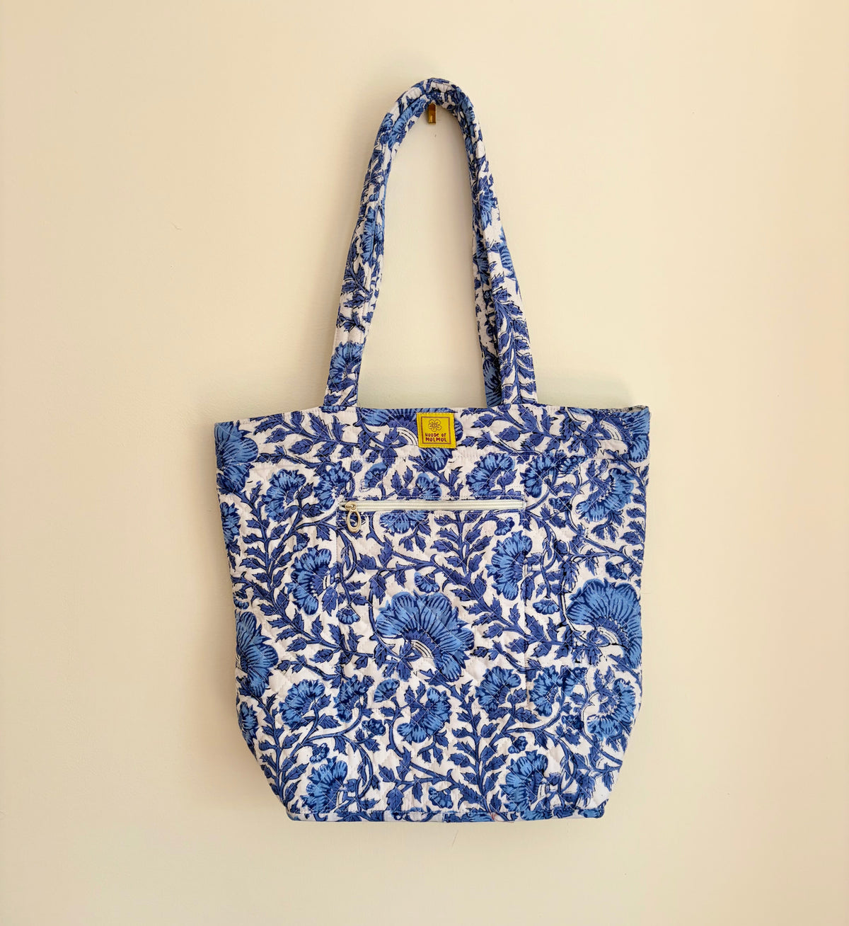House of Mulmul - Bucket Tapered Tote Bag. A blue and white floral print fabric tote bag with two shoulder straps, a front zippered pocket, and a small yellow brand tag, hanging against a plain beige wall.