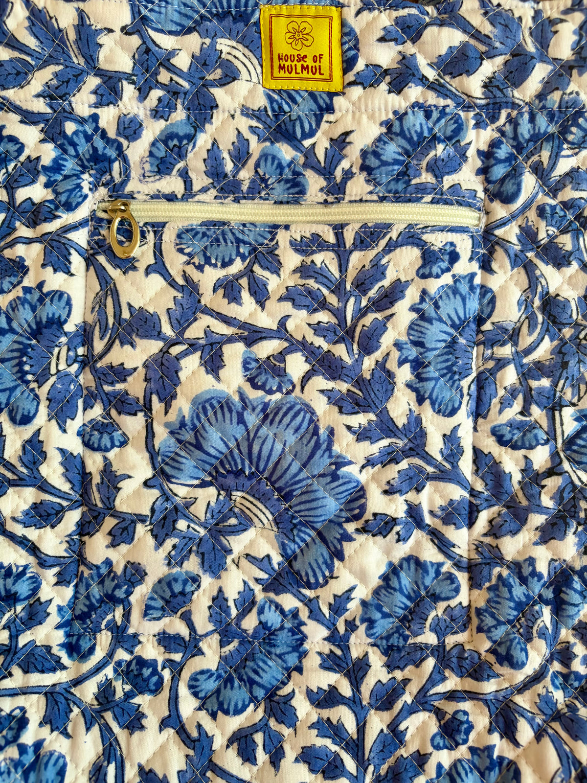 House of Mulmul - Bucket Tapered Tote Bag. Close-up of a quilted fabric with a blue floral pattern on a cream background, featuring a zippered pocket and a yellow label that reads 'House of Mulmul'.