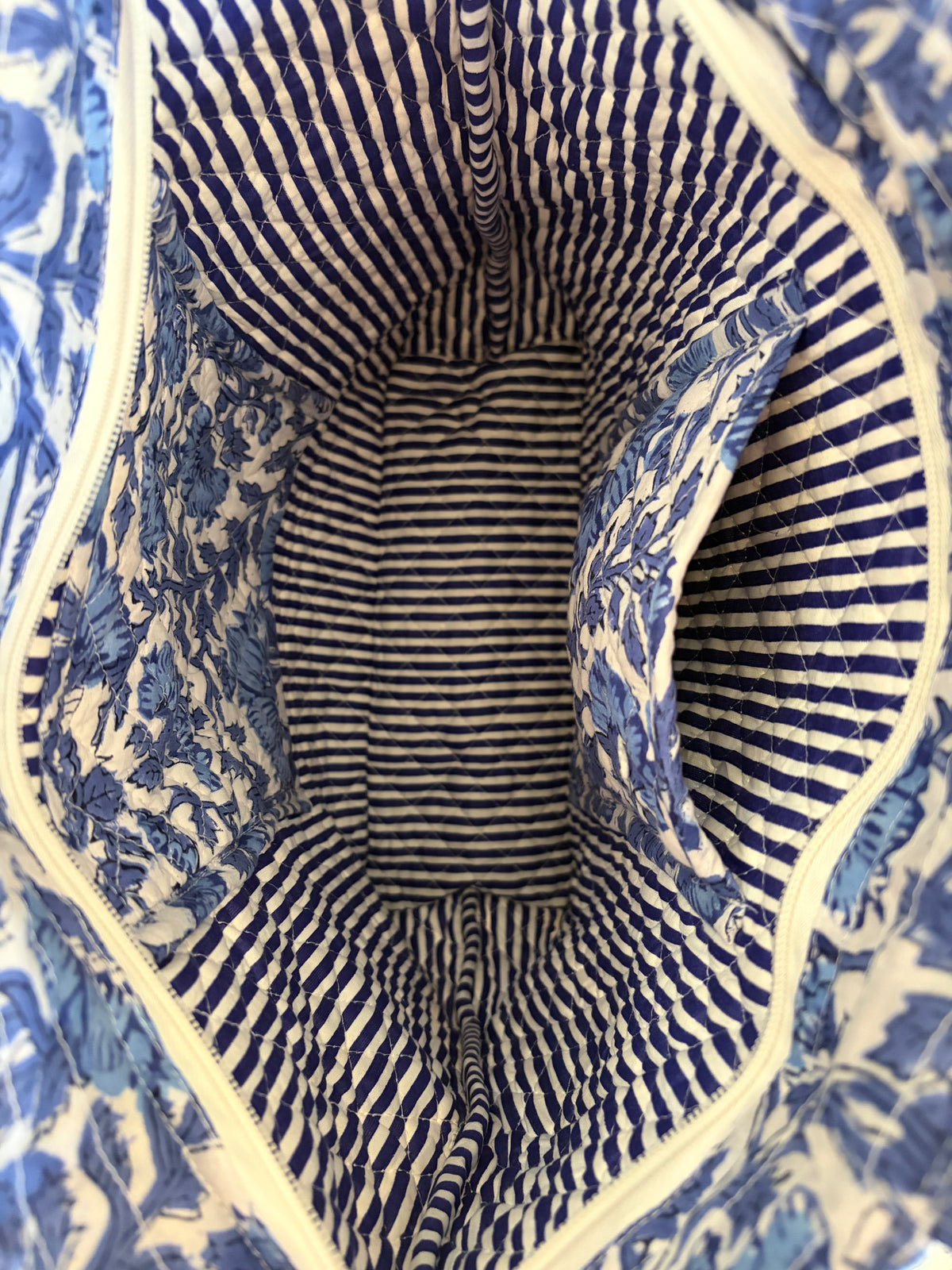 House of Mulmul - Bucket Tapered Tote Bag. Close-up of a quilted tote bag interior, featuring a blue and white floral patterned lining with a striped central pocket and white-colored piping.
