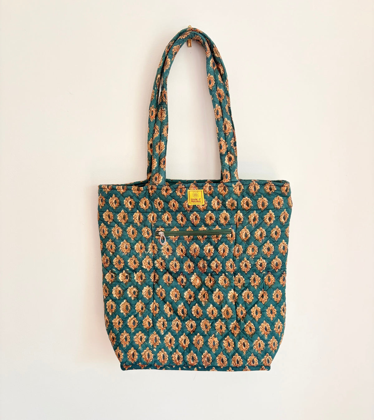House of Mulmul - Bucket Tapered Tote Bag. A fabric tote bag with a teal green and beige floral ikat pattern, featuring dual shoulder straps, a front zippered pocket, and a small yellow brand label.