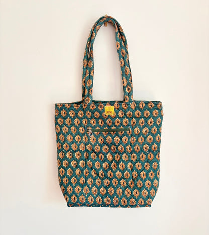 House of Mulmul - Bucket Tapered Tote Bag. A fabric tote bag with a teal green and beige floral ikat pattern, featuring dual shoulder straps, a front zippered pocket, and a small yellow brand label.