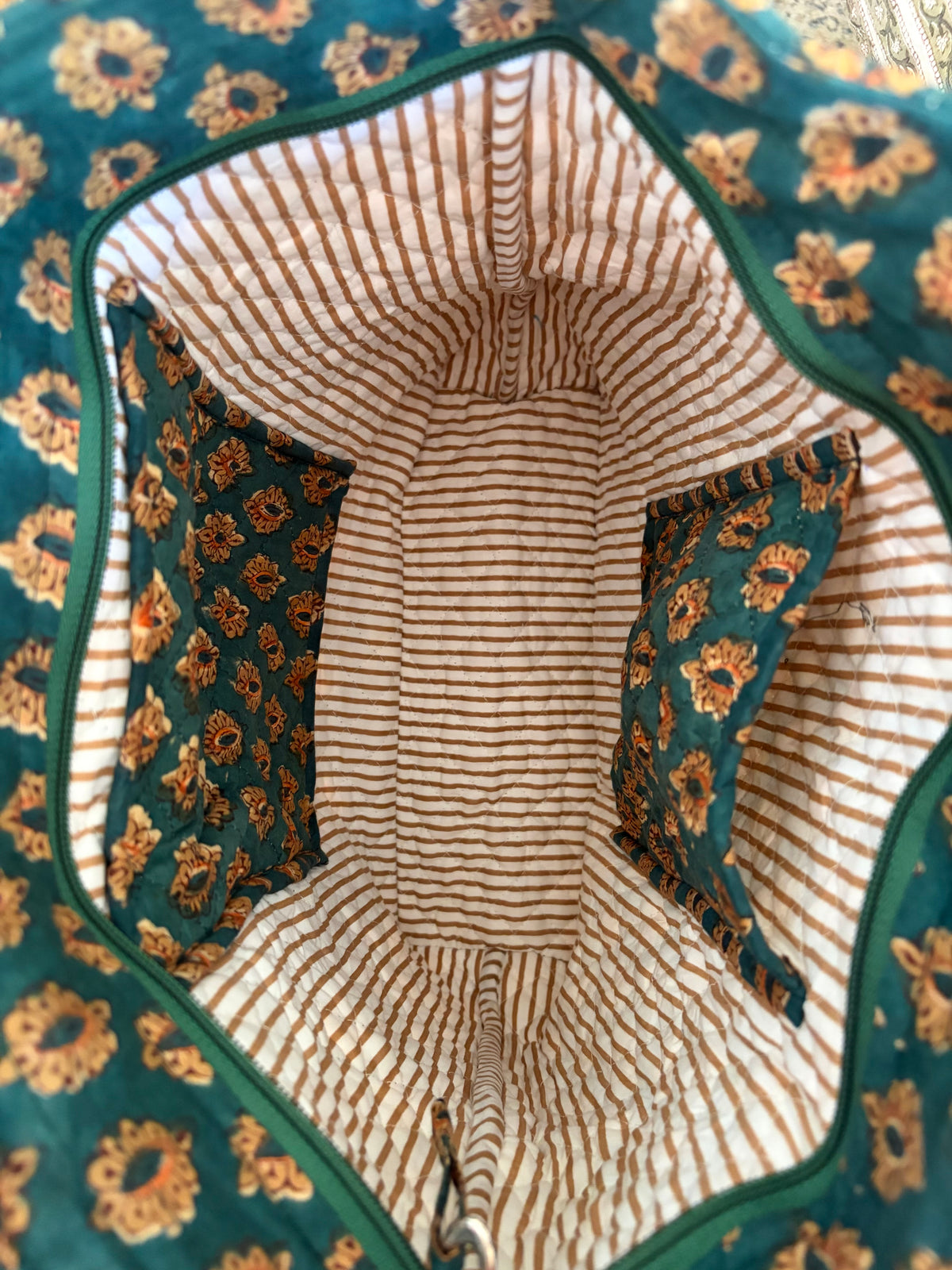 House of Mulmul - Bucket Tapered Tote Bag. Close-up of the interior of a quilted bag or cover, showing a striped cream and brown lining with two teal floral fabric pockets and a matching teal floral outer fabric.
