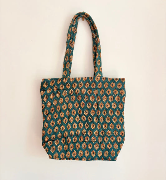 House of Mulmul - Bucket Tapered Tote Bag. A fabric tote bag with a repeating geometric pattern in teal green, brown, and beige, hanging against a plain off-white wall.