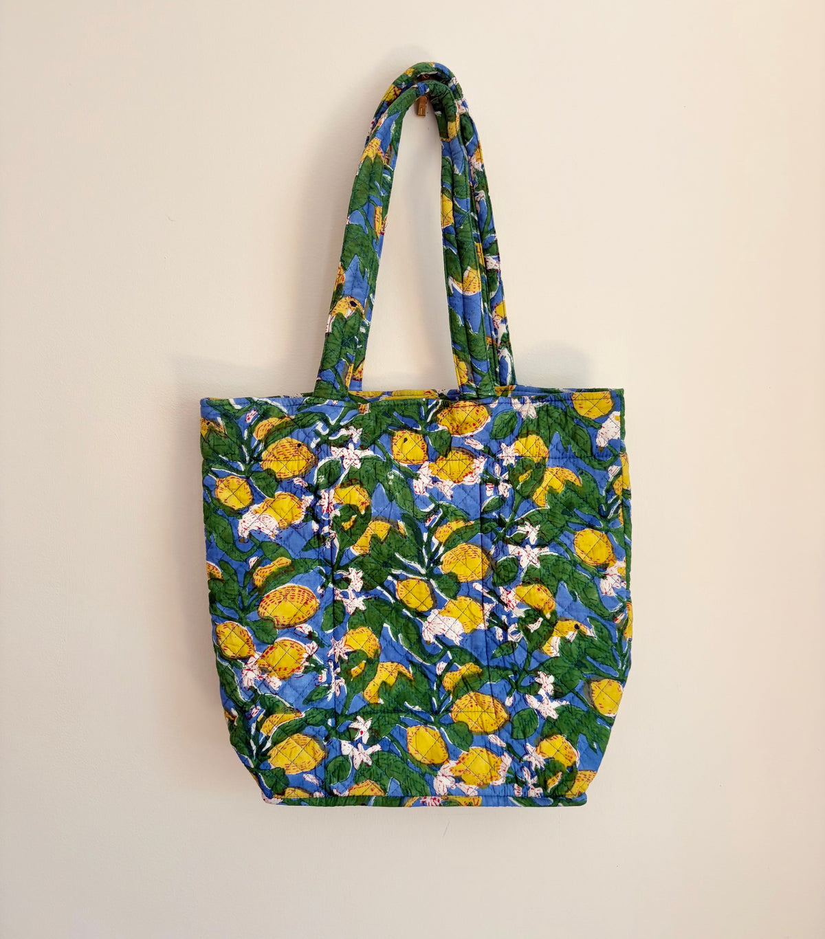 House of Mulmul - Bucket Tapered Tote Bag. A fabric tote bag with a vibrant yellow and white floral print on a blue and green background, hanging against a plain off-white wall.