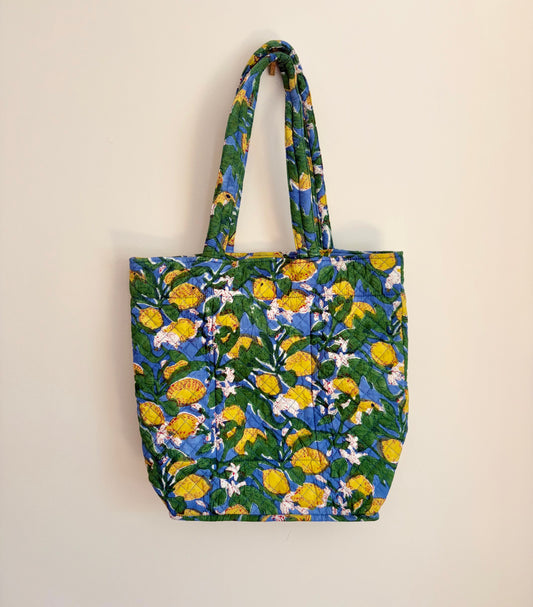 House of Mulmul - Bucket Tapered Tote Bag. A fabric tote bag with a vibrant yellow and white floral print on a blue and green background, hanging against a plain off-white wall.