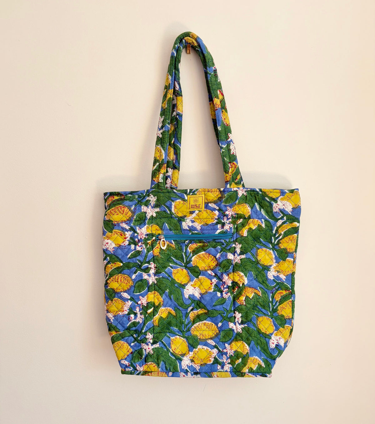 House of Mulmul - Bucket Tapered Tote Bag. A canvas tote bag with a vibrant floral print featuring yellow flowers, green leaves, and blue backgrounds, hanging against a plain off-white wall.