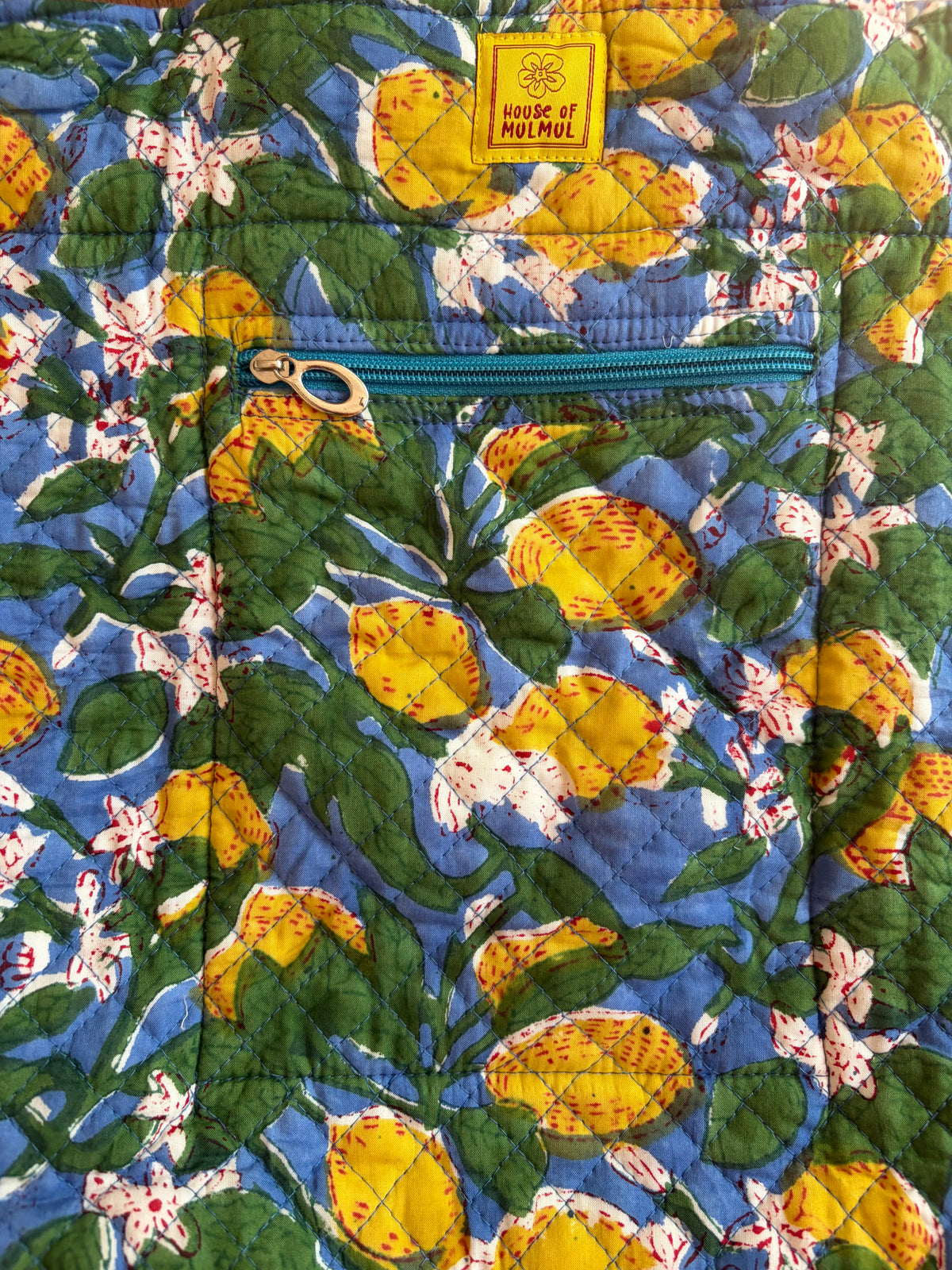 House of Mulmul - Bucket Tapered Tote Bag. Close-up of a quilted fabric item featuring a lemon and white flower print on a blue background, with a yellow 'House of Mulmul' label near the top and a teal zipper pocket.