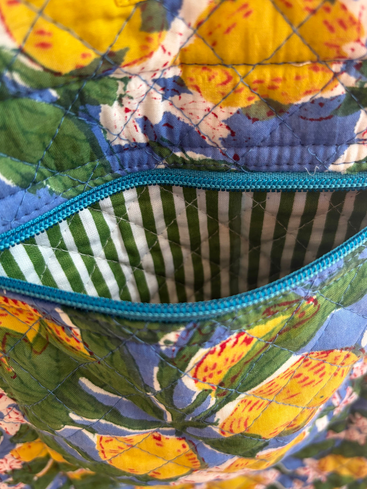 House of Mulmul - Bucket Tapered Tote Bag. Close-up of a quilted fabric bag with a floral print in yellow, green, and blue, featuring a teal zipper and a striped green and white inner lining.