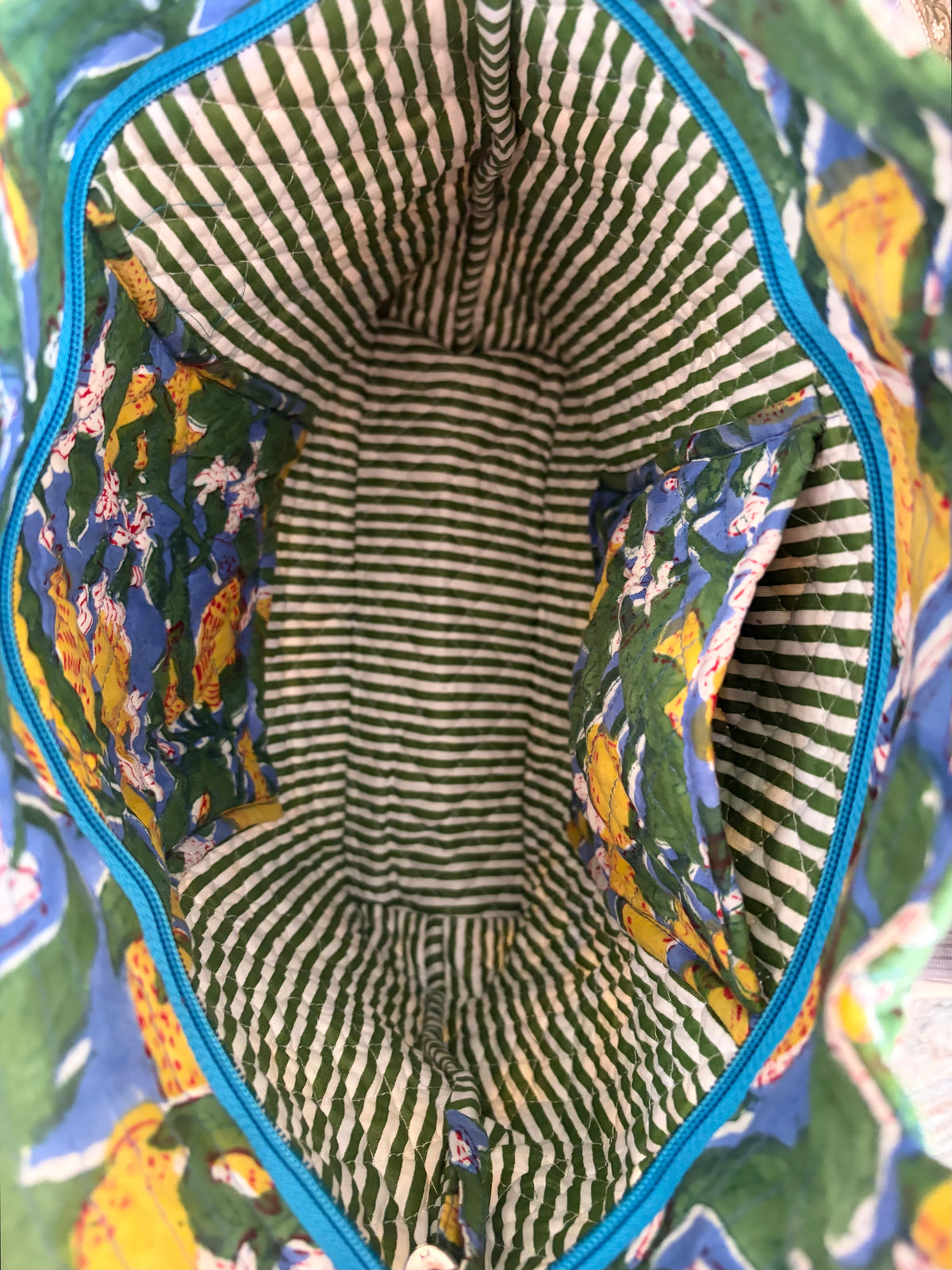 House of Mulmul - Bucket Tapered Tote Bag. Close-up of the interior of a tote bag, showing a striped green and cream fabric lining with floral printed pockets in blue, yellow, and green, framed by a bright blue zipper.