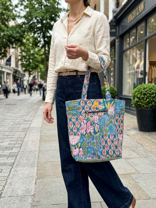 House of Mulmul - Bucket Tapered Tote Bag. A woman on a city sidewalk carries a large tote bag with a colorful floral and butterfly pattern in shades of blue, pink, green, and yellow. She is wearing a cream button-down shirt and dark blue wide-leg jeans, with storefronts and trees in the background.