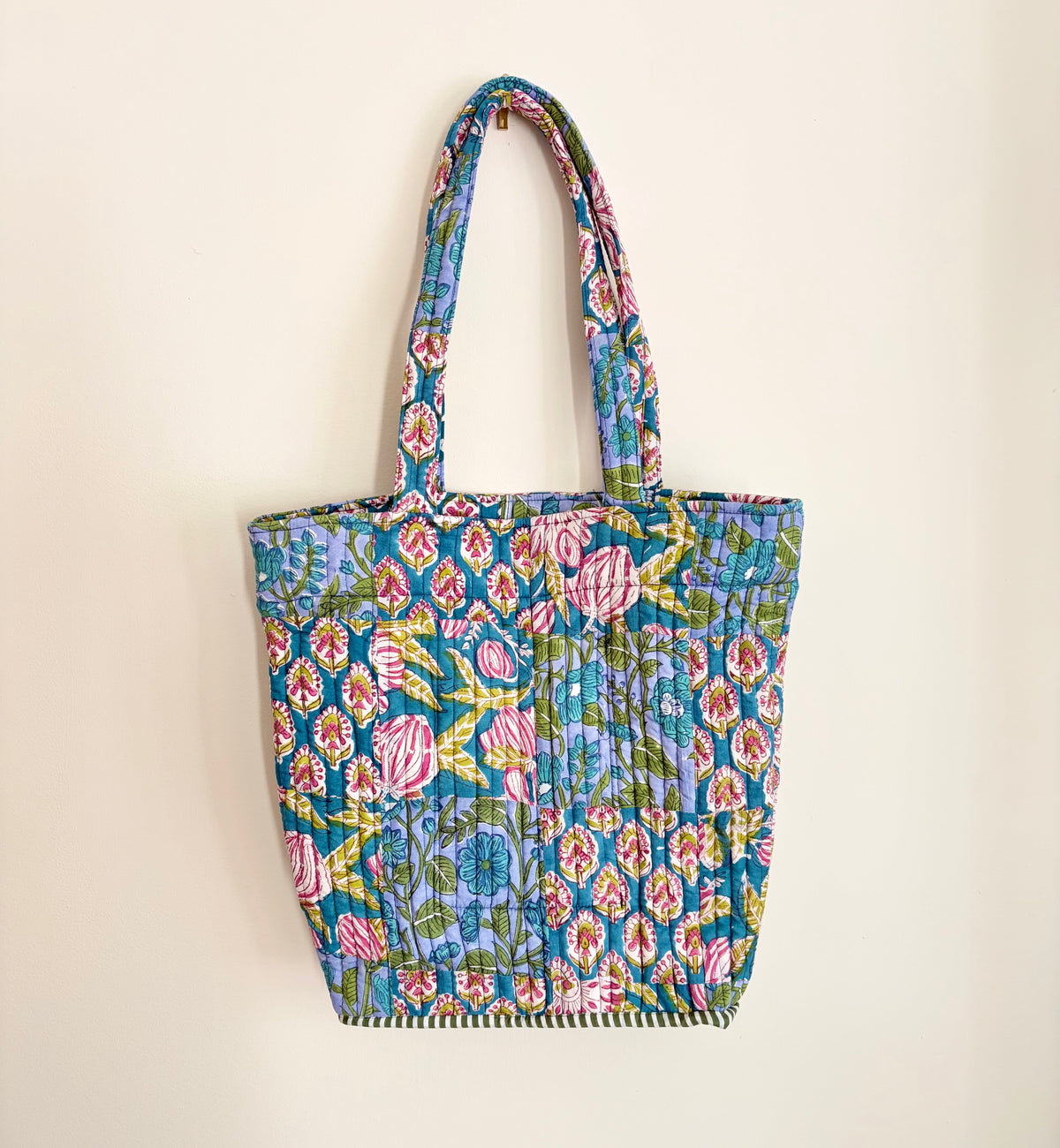 House of Mulmul - Bucket Tapered Tote Bag. A small quilted fabric tote bag with a colorful floral and paisley block-print pattern, featuring two shoulder straps and a gathered bottom hem, shown hanging against a plain off-white wall.