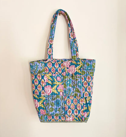 House of Mulmul - Bucket Tapered Tote Bag. A small quilted fabric tote bag with a colorful floral and paisley block-print pattern, featuring two shoulder straps and a gathered bottom hem, shown hanging against a plain off-white wall.