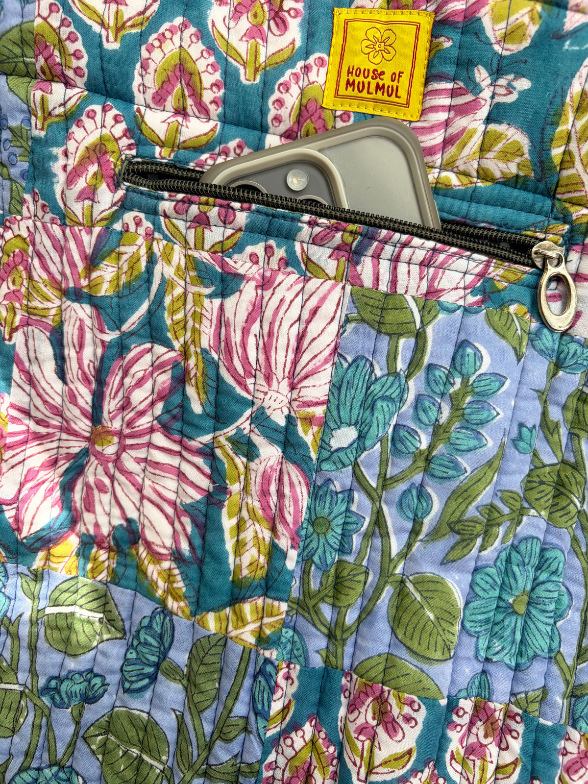House of Mulmul - Bucket Tapered Tote Bag. Close-up of a quilted fabric bag with a floral print in pink, blue, green, and white, featuring a yellow 'HOUSE OF MULMUL' brand patch and a zipper pocket.