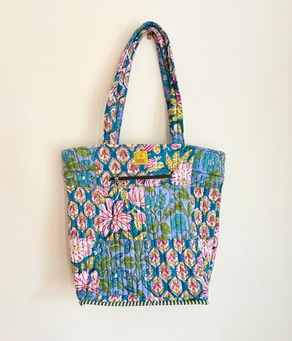 House of Mulmul - Bucket Tapered Tote Bag. A colorful quilted fabric tote bag with a floral and geometric pattern, featuring long shoulder straps and a small yellow brand tag, hanging against a plain off-white wall.