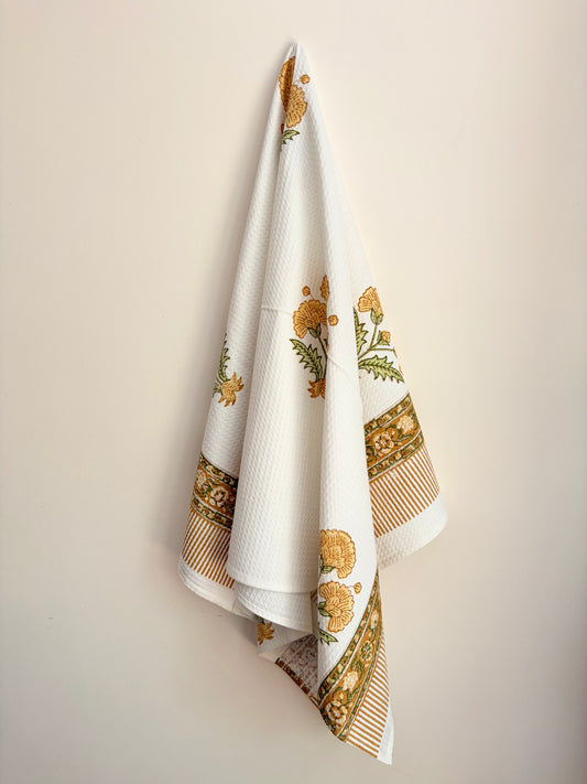 House of Mulmul - Cotton Bath Towel. Decorative white towel with gold and green patterns hanging on a beige wall.