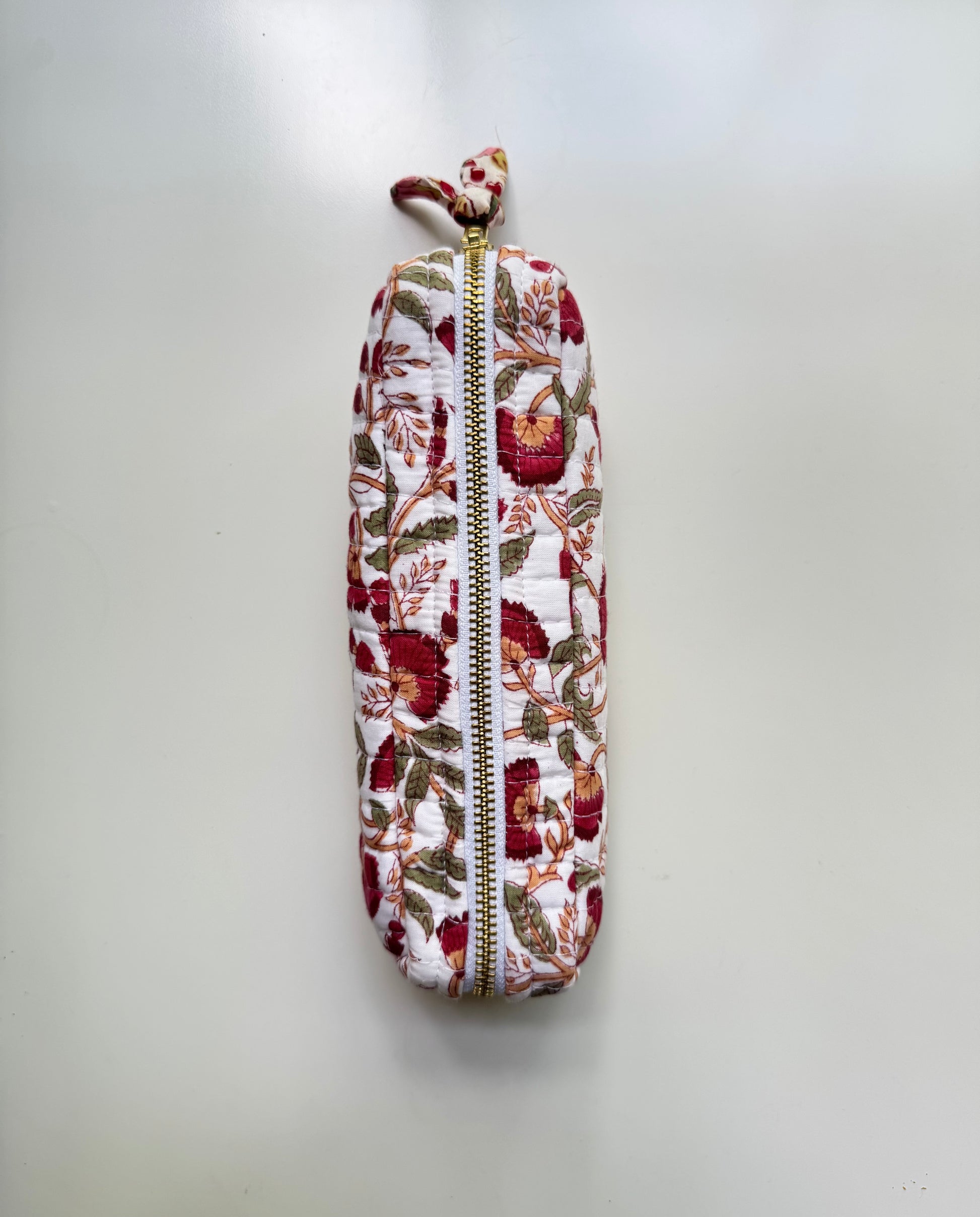 House of Mulmul - Cotton Quilted Makeup Barrel Pouch. A vertical zippered pouch with a white background and a red, green, and brown floral pattern, featuring a small matching fabric loop at the top and a gold zipper.