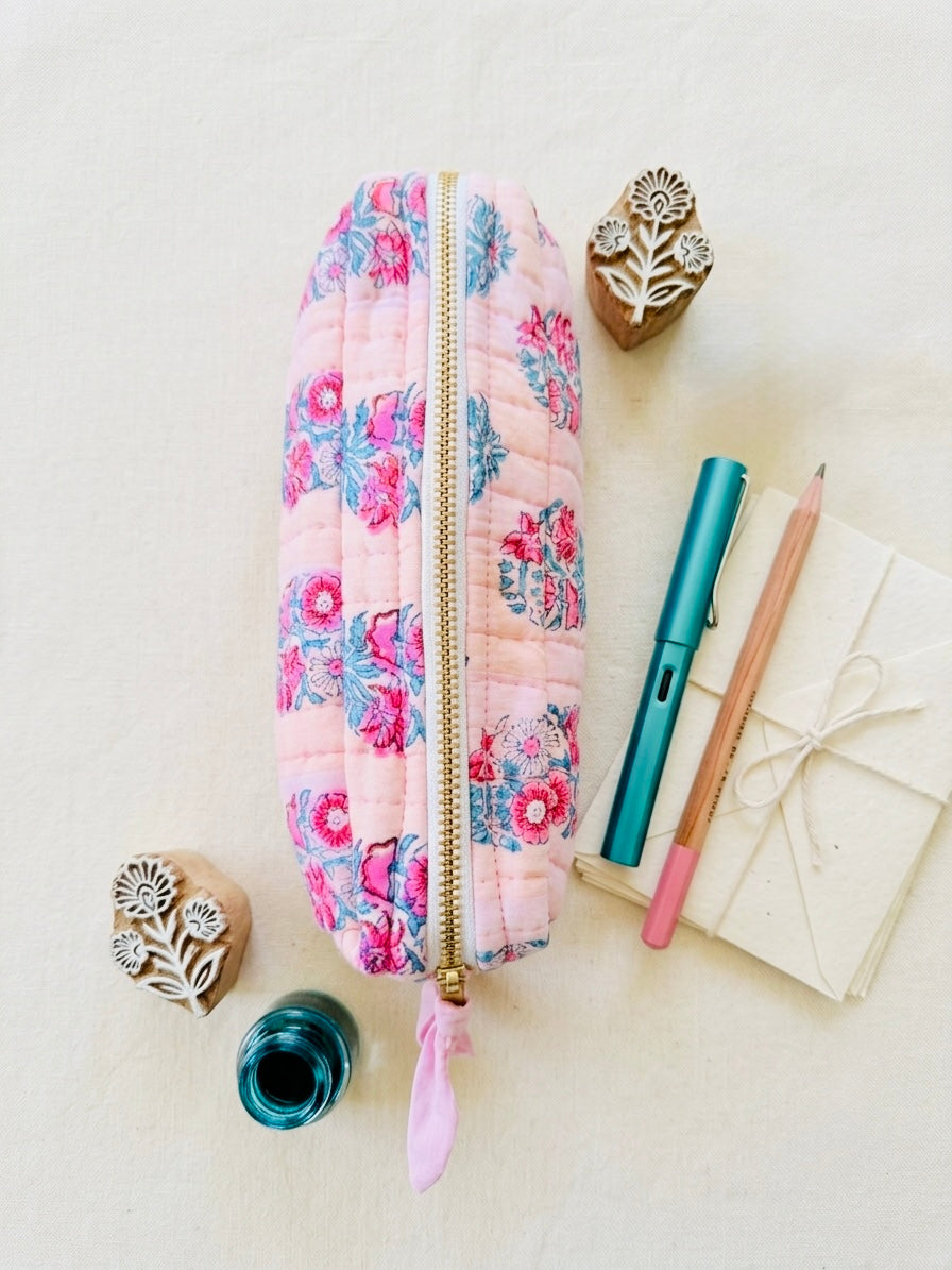 House of Mulmul - Cotton Quilted Makeup Barrel Pouch. A quilted pink zippered pouch with a floral print, styled in a flat lay with stationery and wooden stamp blocks on a light beige background.