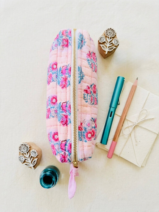 House of Mulmul - Cotton Quilted Makeup Barrel Pouch. A quilted pink zippered pouch with a floral print, styled in a flat lay with stationery and wooden stamp blocks on a light beige background.