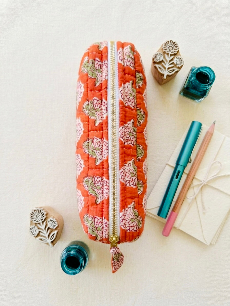 House of Mulmul - Cotton Quilted Makeup Barrel Pouch. An orange quilted fabric pencil case with a pink and green floral block print pattern, styled in a flat-lay arrangement with stationery and rubber stamps on a light cream background.