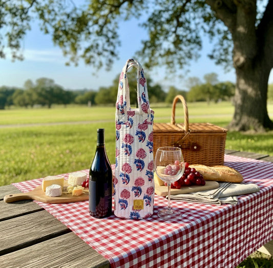 House of Mulmul - Quilted Wine Tote Bag. Picnic setup with a floral bottle cover, wine, cheese, and bread on a checkered tablecloth outdoors.