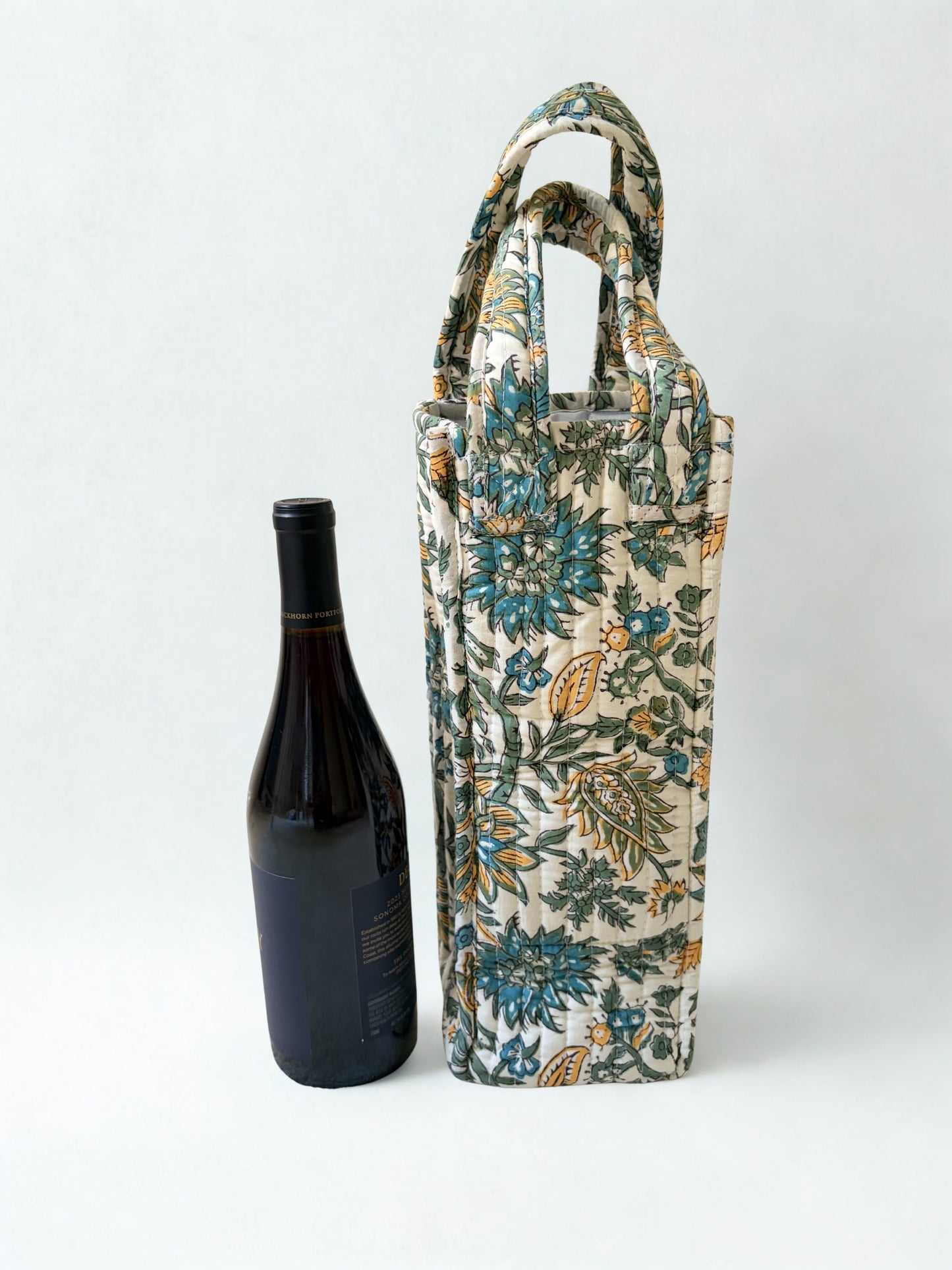 House of Mulmul - Quilted Wine Tote Bag. A tall fabric wine bottle carrier with a floral print in blue, green, and beige, shown next to a dark wine bottle on a light gray background.