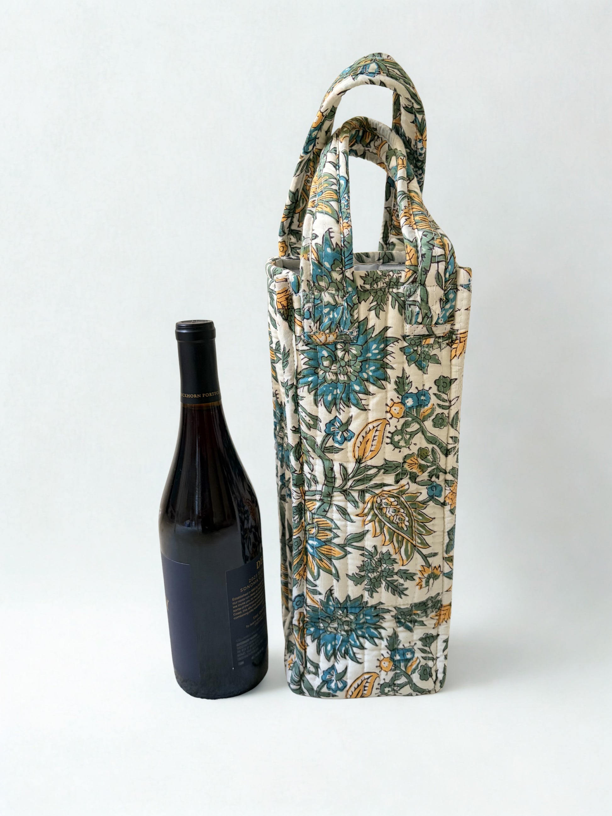 House of Mulmul - Quilted Wine Tote Bag. A tall fabric wine bottle carrier with a floral print in blue, green, and beige, shown next to a dark wine bottle on a light gray background.