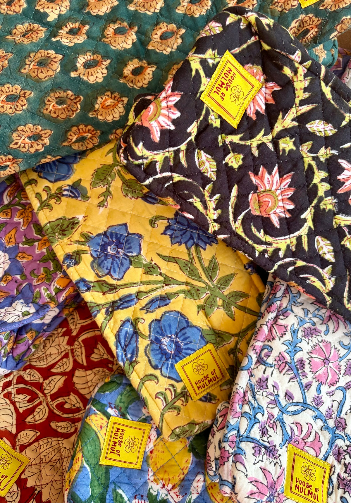 House of Mulmul - Quilted Wine Tote Bags. A close-up of several folded quilted wine bottle bags with colorful floral and botanical patterns, each showing a yellow label that reads 'House of Mulmul'.