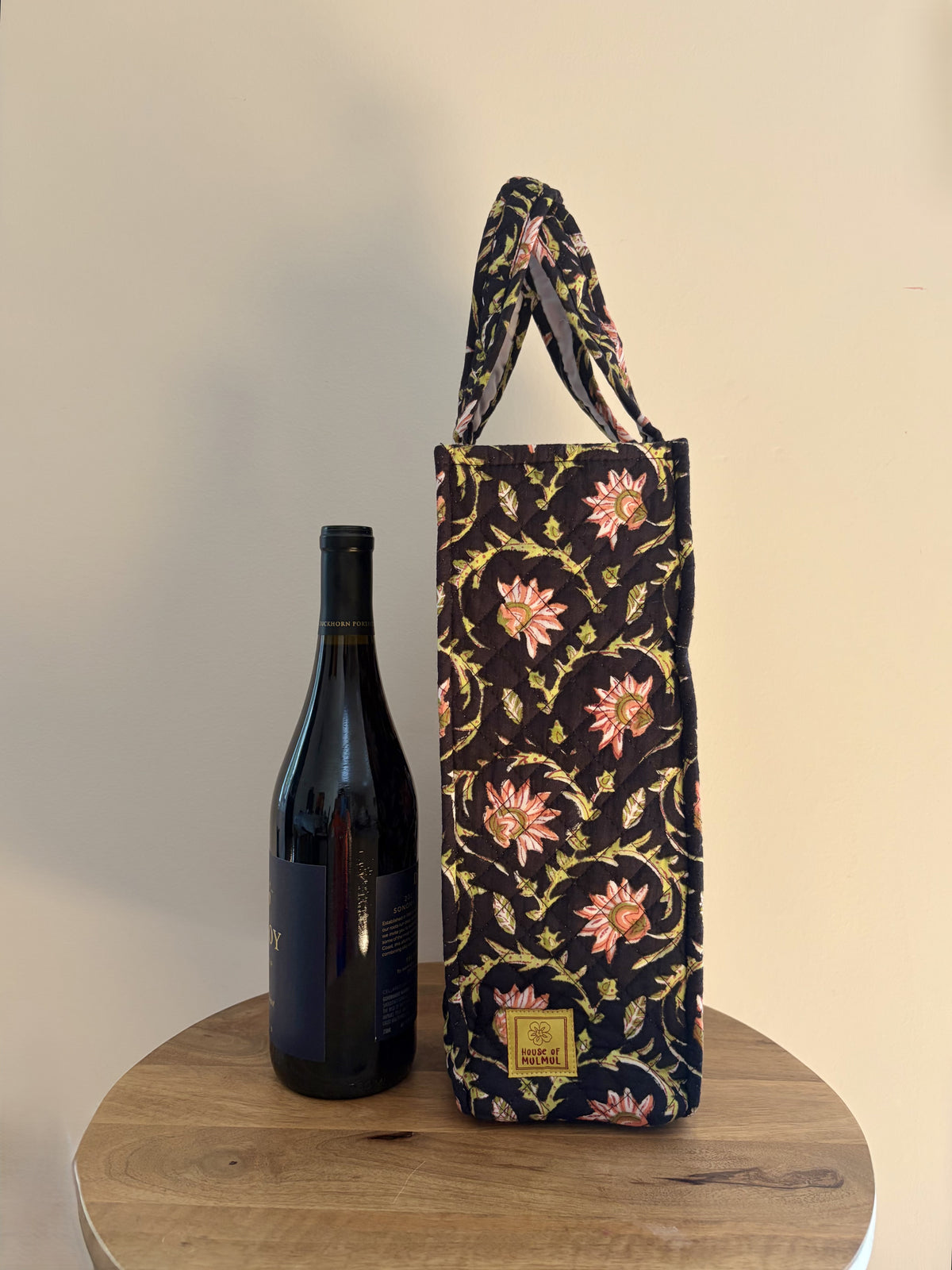 House of Mulmul - Quilted Wine Tote Bags. A floral wine bottle carrier bag with pink and yellow flowers on a dark background, standing next to a dark wine bottle on a round wooden table against a beige wall.