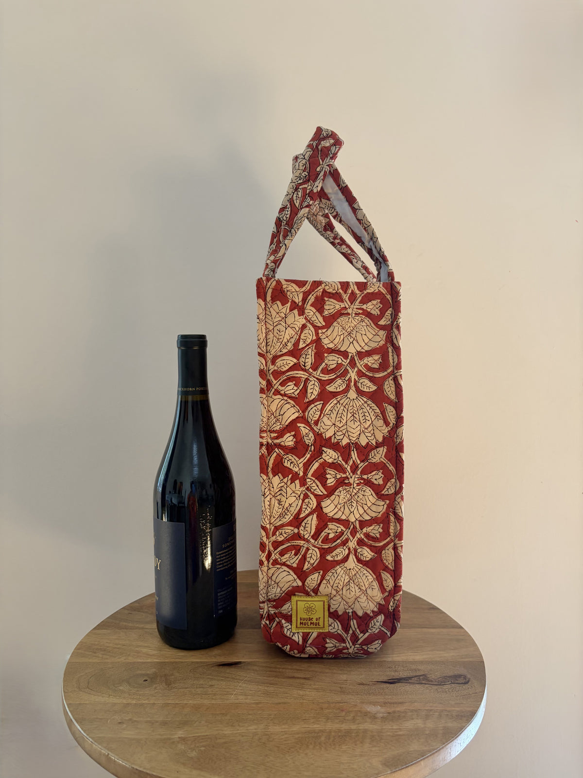 House of Mulmul - Quilted Wine Tote Bags. A red and beige patterned wine bottle carrier with a top handle, displayed on a round wooden table next to a dark wine bottle against a plain beige wall.