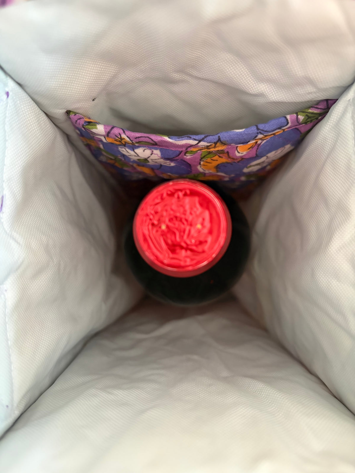 House of Mulmul - Quilted Wine Tote Bags. A top-down view of a dark bottle with a bright red cap nestled inside a white insulated bag or cooler, with a colorful floral fabric pocket visible behind it.