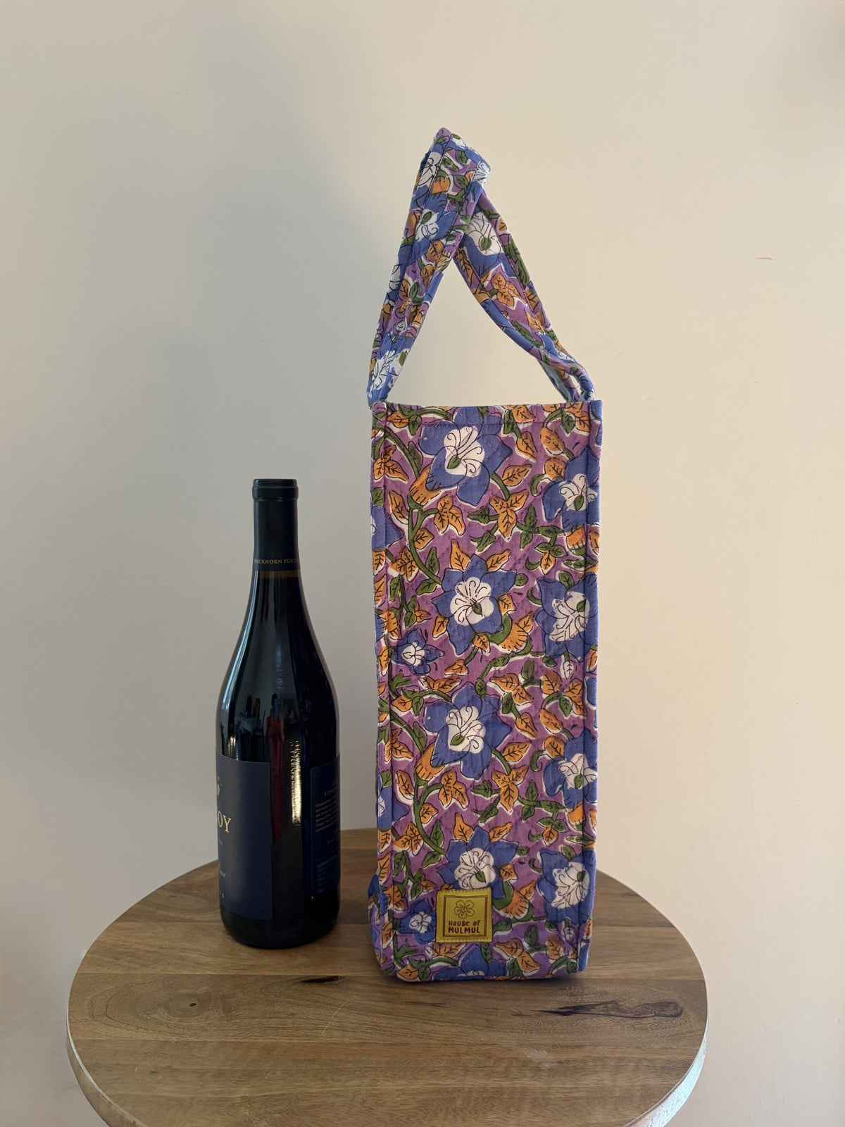 House of Mulmul - Quilted Wine Tote Bags. A floral wine bottle carrier bag with a purple base and multicolor print, shown next to a dark wine bottle on a round wooden table against a plain beige wall.